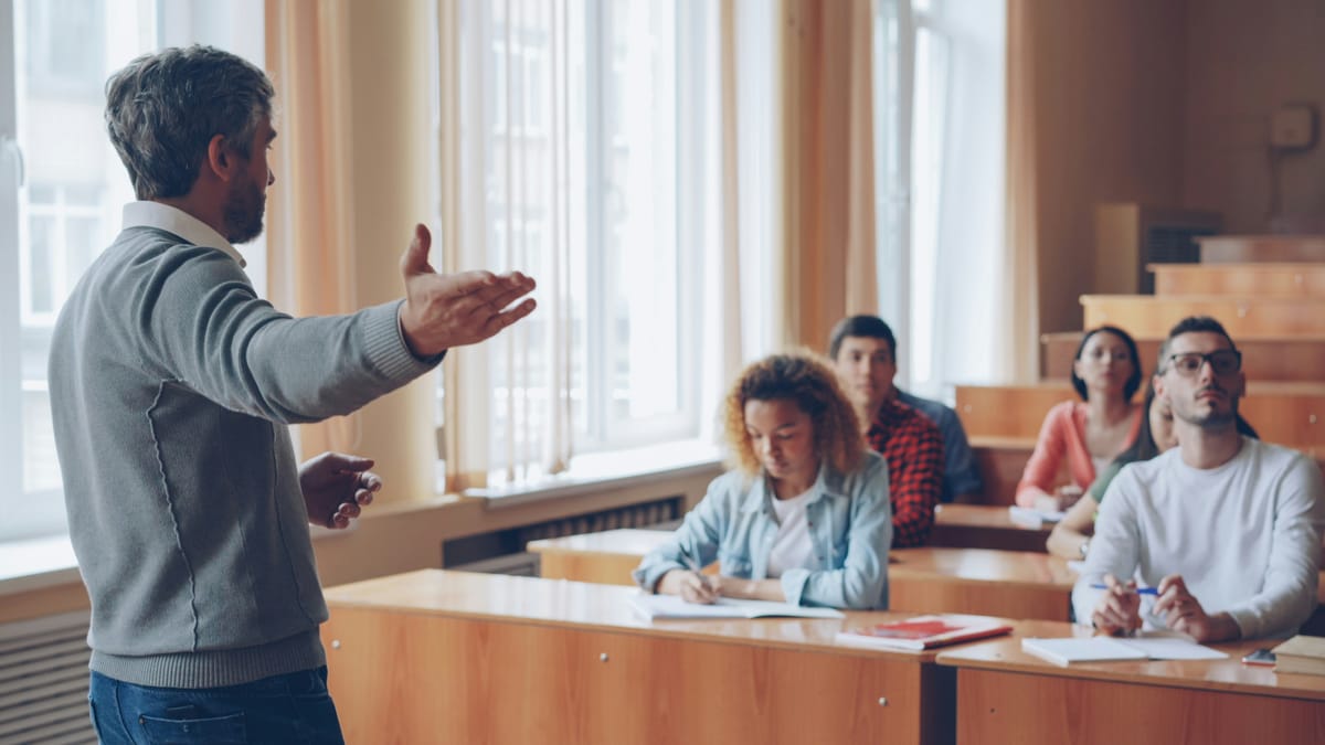 A teacher gesturing as adult students at their desks listen and take notes.
